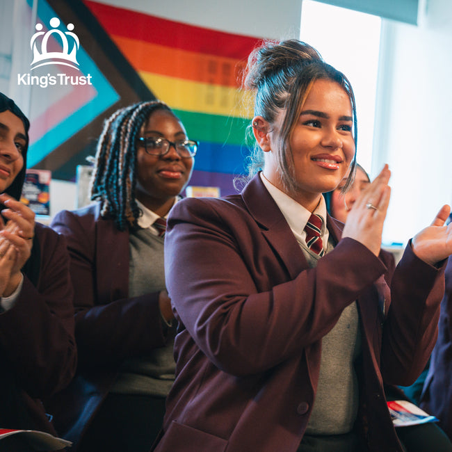 Students in school uniforms clapping and smiling during a classroom event, with a Progress Pride flag in the background. The image includes the King’s Trust logo, representing a partnership with Tropic to support young people through education and empowerment programs.